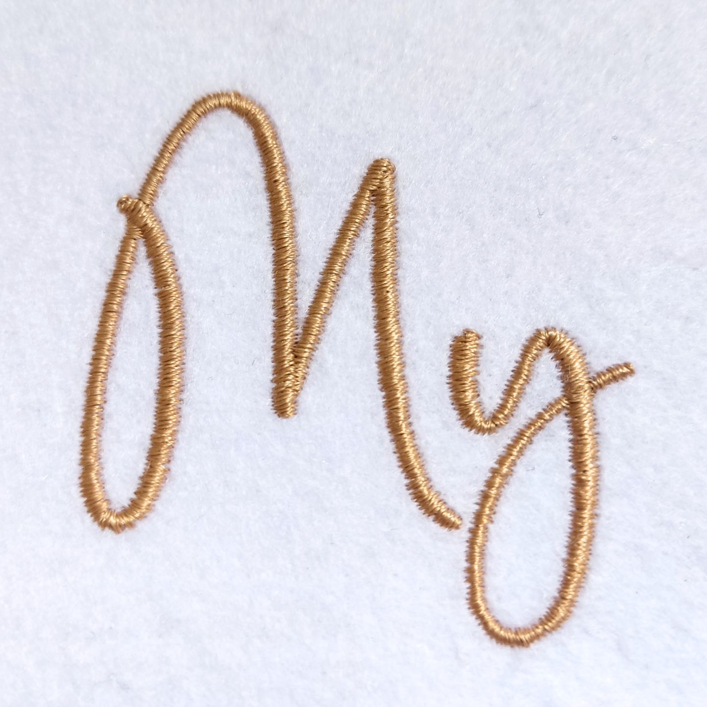 Gold embroidered cursive lettering spelling 'My' on a white textured fabric background.