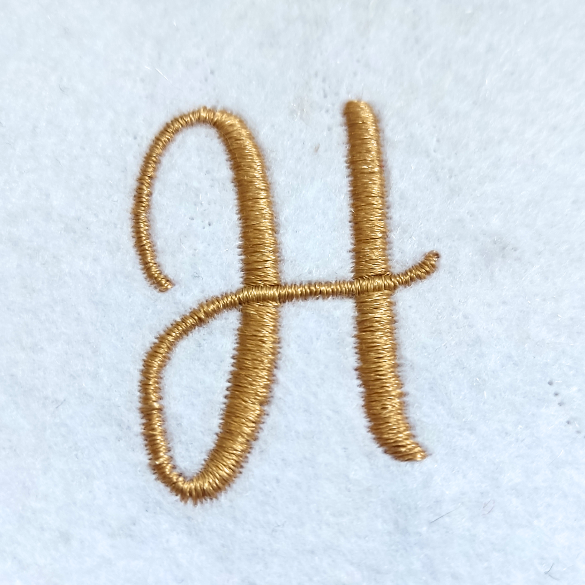 A close-up of a gold embroidered cursive letter 'H' on a white textured fabric background.
