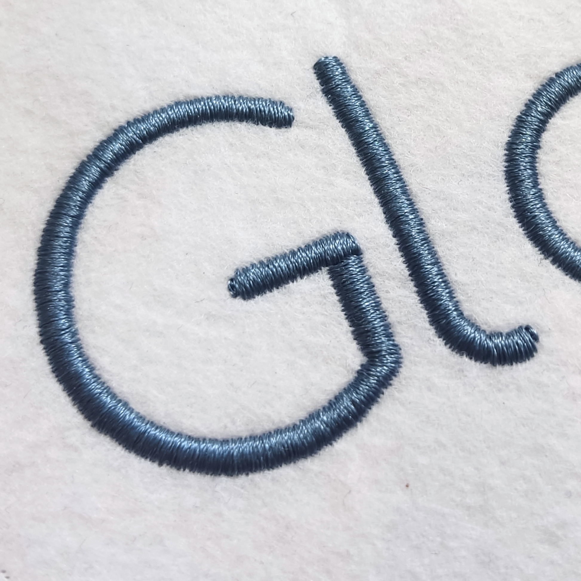 Close-up of embroidered lettering in a dark blue-gray thread on a white fabric background.