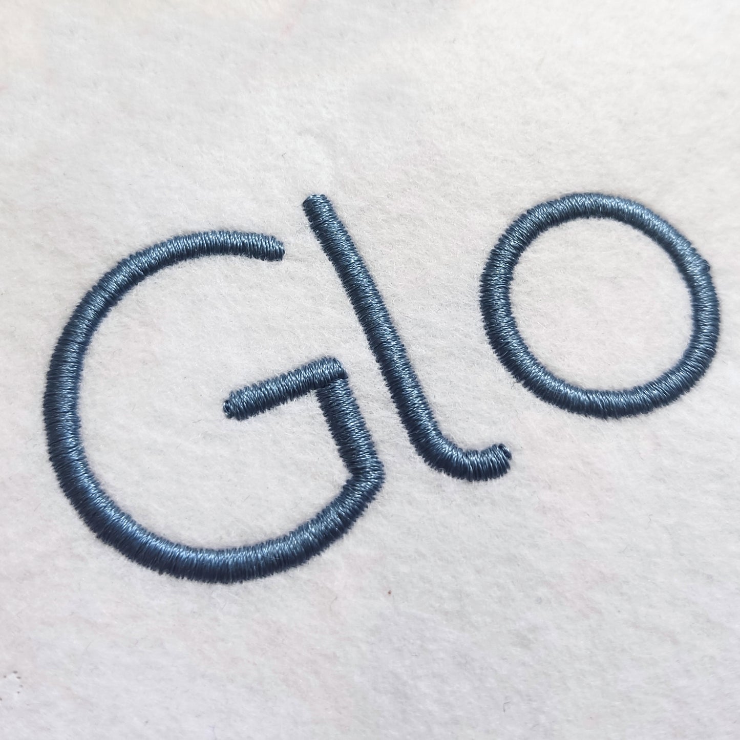 Close-up of embroidered lettering on a white fabric surface, showing the partial word 'Glo' in a muted blue-gray thread.