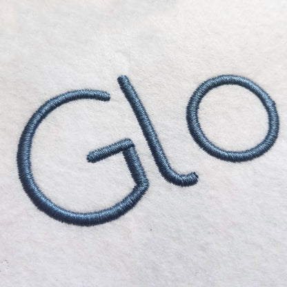 Close-up of embroidered lettering on a white fabric surface, showing the partial word 'Glo' in a muted blue-gray thread.